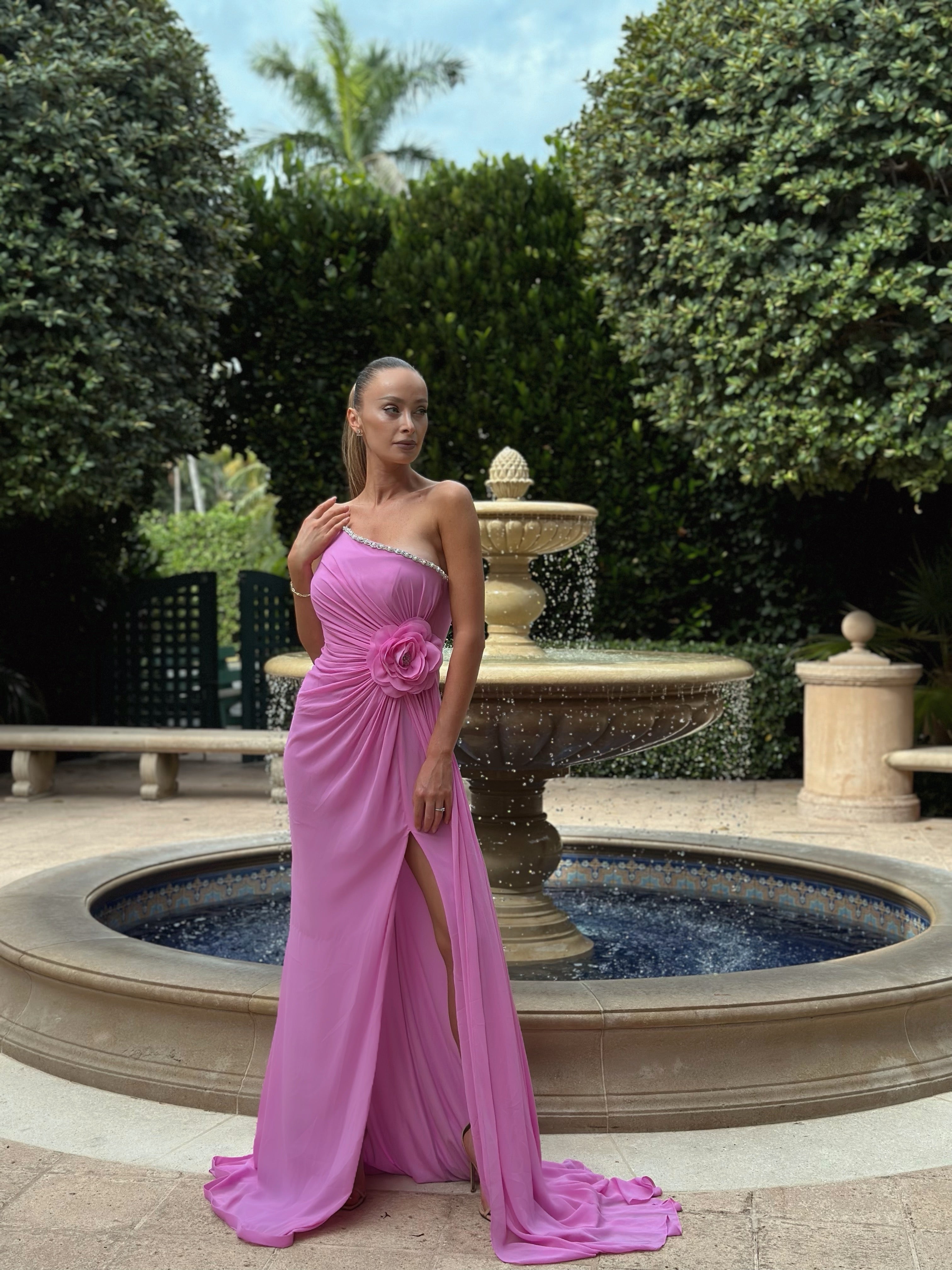 Elegant pink one-shoulder gown with floral embellishment at the waist and a high slit, perfect for formal events and evening occasions