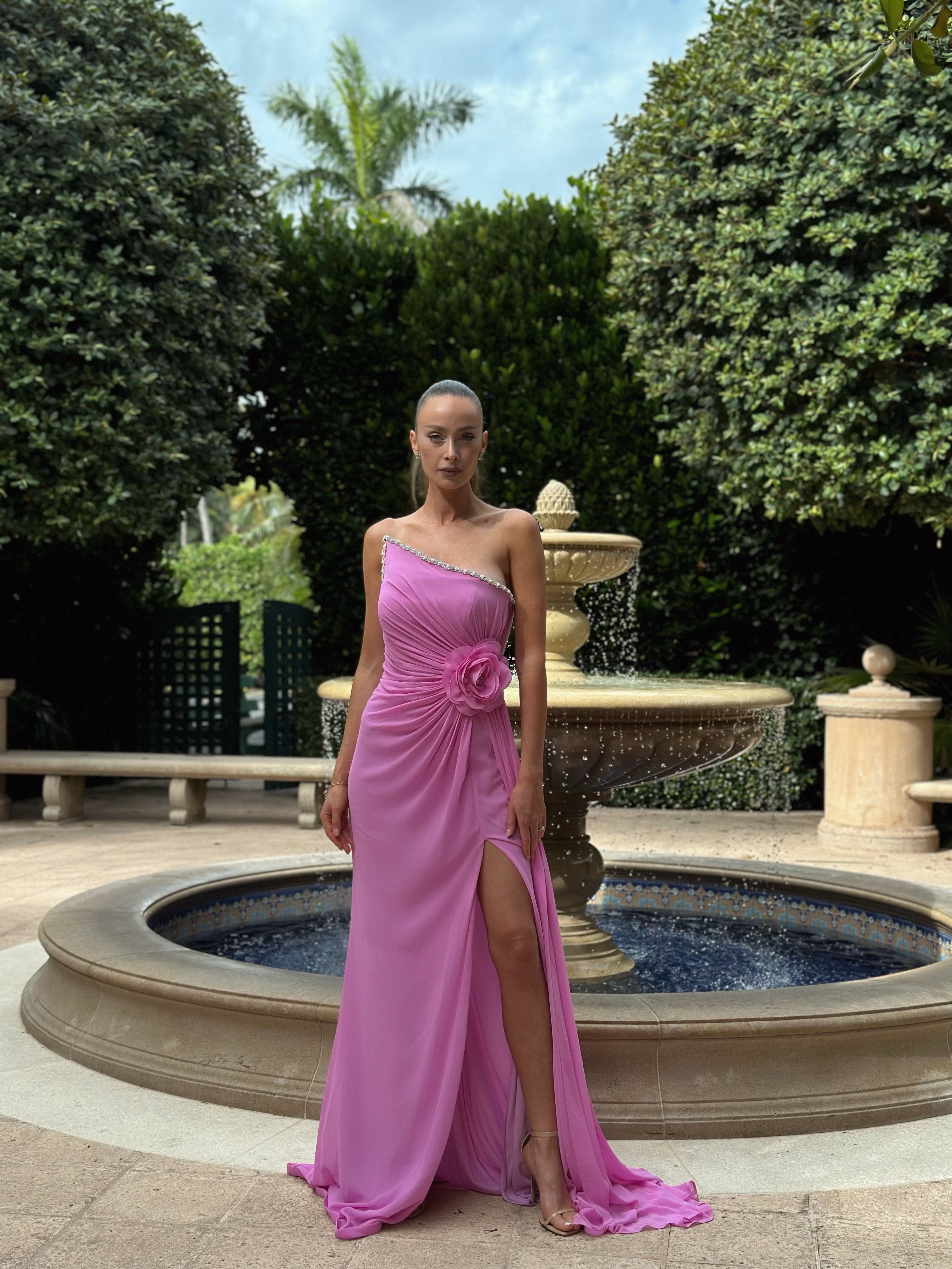 Elegant pink one-shoulder gown with floral embellishment at the waist and a high slit, perfect for formal events and evening occasions