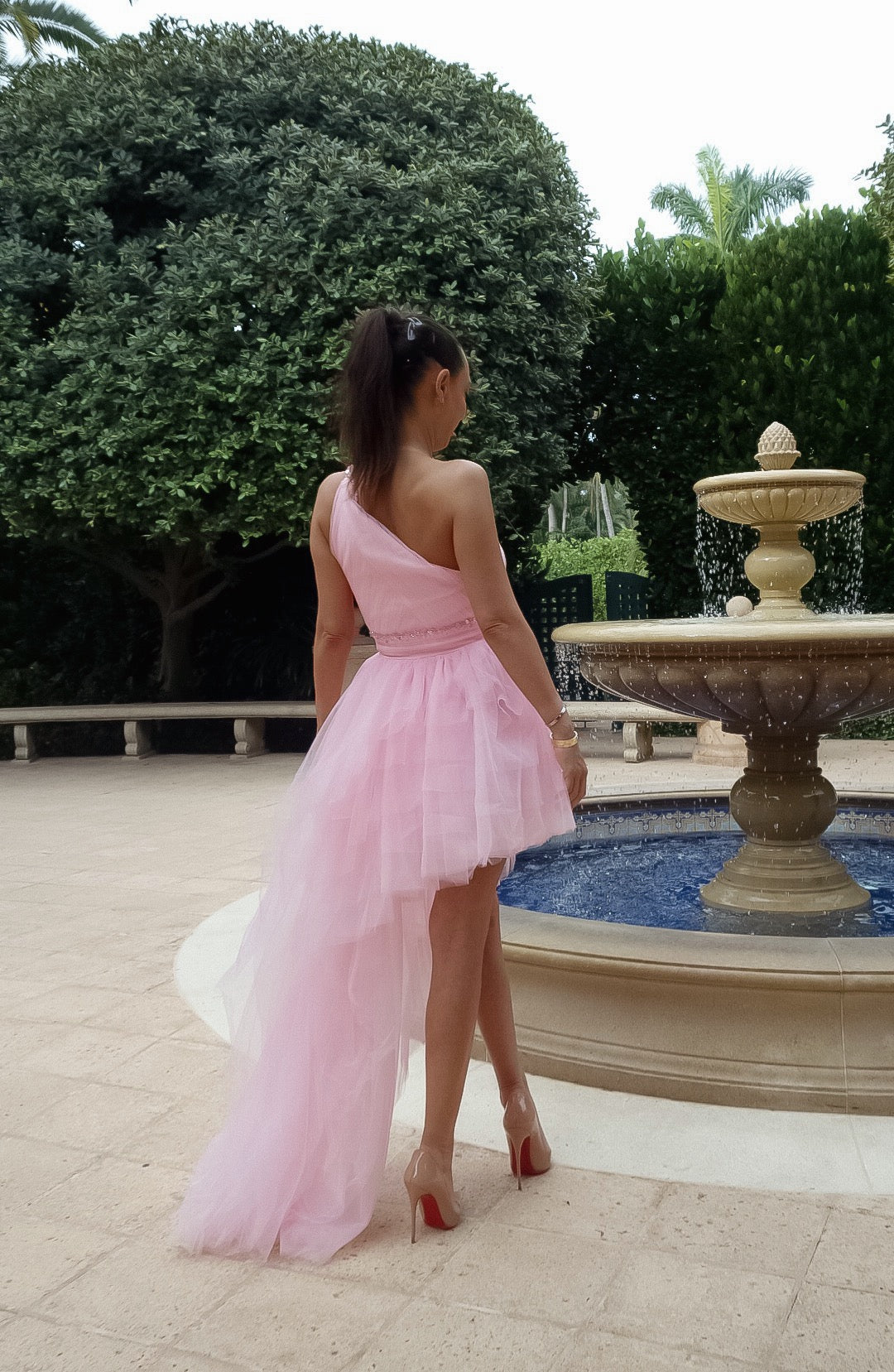 One-shoulder pink dress made from soft tulle with a high-low hemline, trailing back for a whimsical effect, and cinched waist with a subtle belt, perfect for special events.